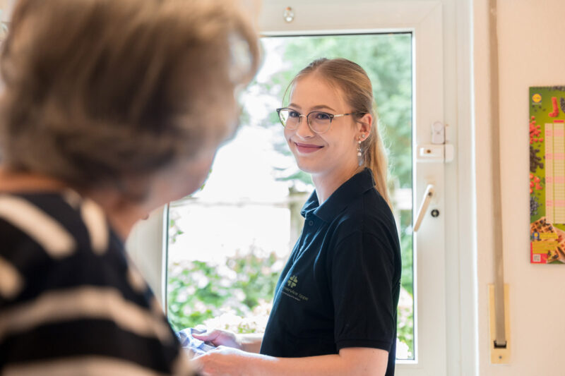Szene: Mitarbeiterin des Seniorenservice lächelt bei Reinigungsarbeiten in der Wohnung einer Klientin in die Kamera