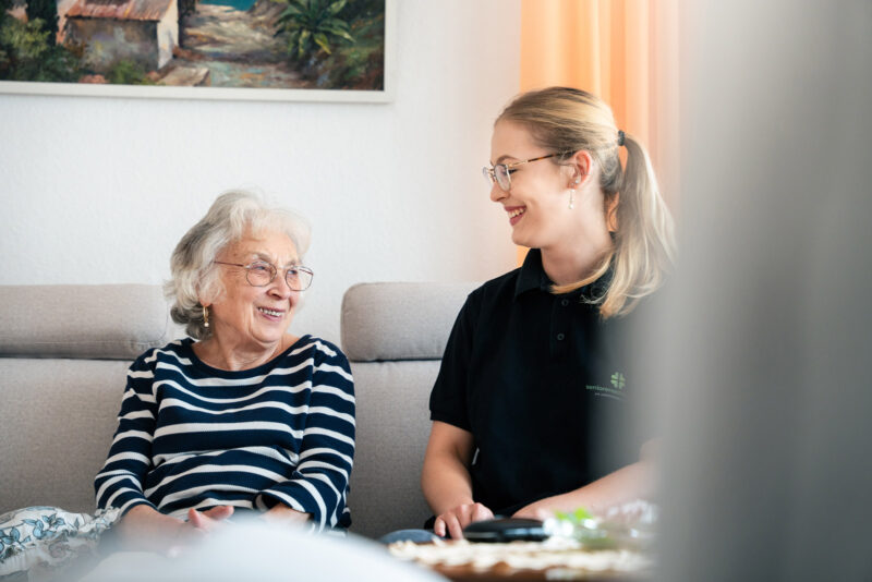 Szene: Mitarbeiterin des Seniorenservice mit einer älteren Dame auf dem Sofa