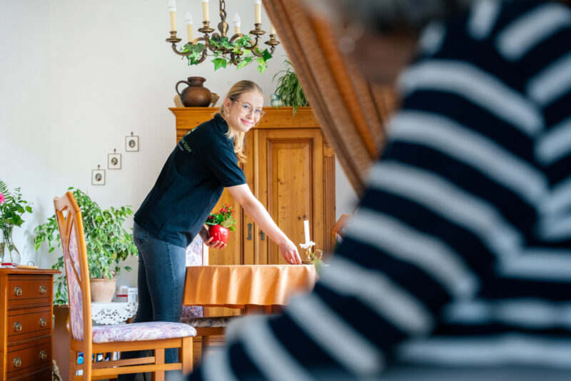 Szene: Lächelnde Mitarbeiterin beim Aufräumen in der Wohnung einer Klientin